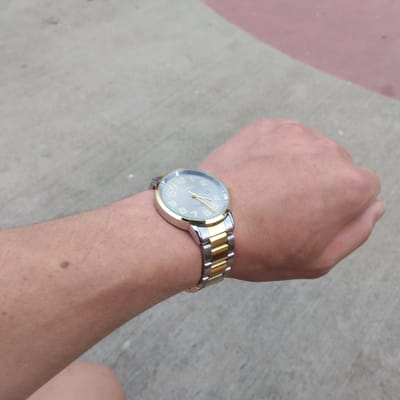 A person's left forearm and wrist, adorned with a two-tone metal watch, are captured in a close-up, outdoor shot. The watch features a silver-colored band with gold-colored accents and a grey dial with gold-colored hour markers and hands. The forearm displays light skin with visible hair and a small, dark mole. The background consists of a textured grey surface, likely concrete or asphalt, with a diagonal band of reddish-pink material on the right side, suggesting an outdoor urban setting possibly in Pilsen, Czechia. The lighting indicates it is daytime.