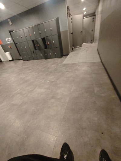 A gray-toned locker room or changing area. Lockers line the left wall. A door with a small window is visible in the background, hinting at a bathroom or shower area. The floor is gray tiled. In the foreground, the lower part of the view shows the person's legs and shoes. The lighting is muted and even, highlighting the utilitarian nature of the space. The room is empty.