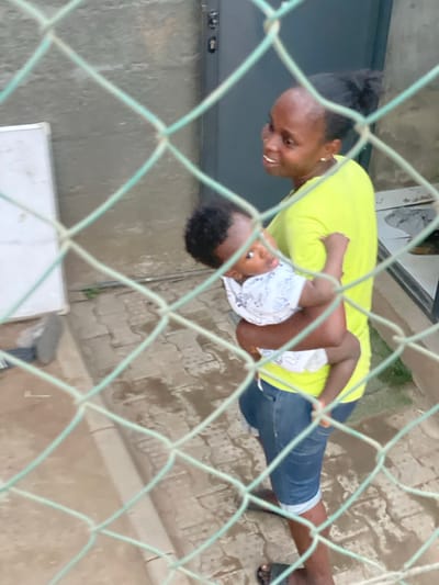 A woman in a neon yellow shirt and denim capri pants holds a baby wearing a patterned white and blue shirt. The woman has dark hair pulled back and is looking down at the baby. Both are dark-skinned. The image is taken through a green wire fence, slightly obscuring the subjects. The background is a paved area and part of a building with a door.
