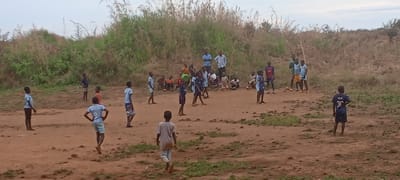 The image captures an outdoor scene in Ngalula, Democratic Republic of the Congo, featuring a large group of people, predominantly children, gathered on a dirt field. The setting appears rural, with sparse vegetation and tall grasses lining the background, suggesting an undeveloped area.
The primary activity seems to be an informal game of football (soccer) or a similar group play. Many children are positioned on the field, some in stances suggestive of active play, while others are standing or observing. Several boys are wearing football jerseys, including one on the far right with the number "30" visible on his dark blue shirt. Another boy in a light blue shirt in the foreground has an emblem or text on his back, but it's indistinct.
In the background, a cluster of children sits or stands, watching the activity. A few adults are also present, standing further back, possibly supervising or observing the game. The ground is uneven, reddish-brown dirt, with patches of green grass. The sky is overcast, indicating it is daytime, possibly late afternoon. The overall atmosphere conveys a sense of community and children engaged in recreational play in their natural environment.