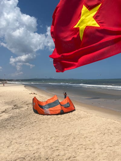 The image shows a beach scene in Phu Thuy Ward, Vietnam, with a red flag bearing a yellow star prominently displayed in the foreground. The flag is partially obscuring the view of the sky, which is a clear blue with scattered white clouds.

In the middle ground, an orange and grey kite is laid out on the sand, with a person visible behind it, seemingly preparing for a water sport. The beach itself is sandy and appears to be quite wide, with gentle waves breaking on the shore. In the distance, there are multiple kites in the air, indicating a kitesurfing or kiteboarding event is taking place. Several people can be seen scattered along the shoreline and in the water.

The time of day appears to be daytime, likely late morning or early afternoon, given the brightness of the sunlight. The weather seems pleasant with a clear sky and moderate waves. The presence of the Vietnamese flag suggests a connection to Vietnam, and the location context confirms it as Phu Thuy Ward.

The kite has visible text that reads "FLY SURFING" and "STOP 9". The overall mood of the image is active and recreational, suggesting a sunny day with opportunities for water sports.