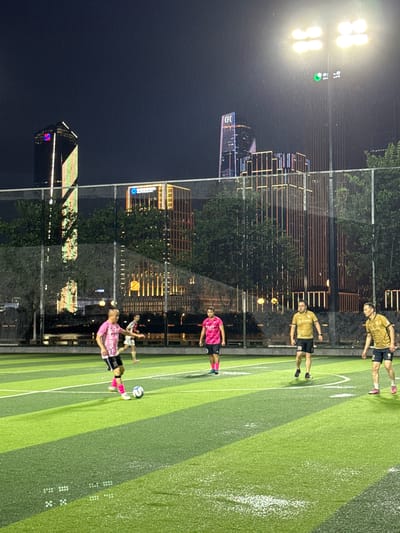 In Haizhu District, China, at night, several individuals are playing soccer on an illuminated artificial turf field, likely a sports complex or park, as indicated by the presence of floodlights. The backdrop features a cityscape with tall, modern buildings illuminated with colorful lights. Three individuals in yellow soccer jerseys and black shorts are on the field, along with two individuals in pink jerseys and black shorts. One person in a pink jersey is actively kicking a soccer ball. The scene is set against a dark sky, with visible light rain or water droplets in the air.