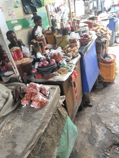 The image depicts a bustling market scene in Alaiyabiagba, Nigeria. Two women are seated behind stalls laden with various food items. One woman is observed eating, while the other is looking at a smartphone while wearing earphones. The stalls display a variety of goods including peanuts in a red tin, onions, various snacks in plastic bags, and other produce in containers. The environment is characterized by a somewhat crowded and gritty setting, with the ground appearing worn and stained. A blue tarp covers part of one of the stalls, and various buckets and containers are scattered around. Bags of what appear to be dried peppers are placed in the foreground.