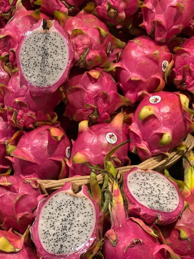 A close-up, overhead shot captures a vibrant display of dragon fruits, both whole and halved, piled within a rustic woven basket. The fruits exhibit their characteristic bright pink skin, some with greenish tips on their leathery scales, and some are sliced open to reveal their white flesh speckled with tiny black seeds. The lighting highlights the glossy sheen of the fruit skins. The image appears to be taken in a market setting, potentially at the Warorot Market (Kad Luang) in Chiang Mai City Municipality, Thailand, known for its abundant fresh produce. No people are visible in the frame.