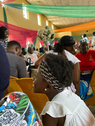 The image shows a gathering of people inside a hall decorated with draped fabric in shades of green, red, and orange. The hall appears to be a place of worship or a community event space. The lighting suggests it could be daytime, with natural light filtering in from windows visible at the top of the walls, or artificial lighting is in use.
In the foreground, a woman is visible from the side, wearing a white dress with ruffled sleeves and a black and white patterned headscarf. She has an earring in her visible ear. To her left, a portion of another person's lap is visible, covered by a colorful fabric featuring comic book style illustrations.
In the middle ground, several people are seated, some facing away from the camera and others in profile. A man in a grey shirt is seated to the left. Further back, a woman in a white outfit with red patterned fabric is seated, and to her right, a man in a red shirt and a blue and white patterned cap is looking towards the right.
In the background, more people are visible, some standing and some seated. A large floral arrangement of white flowers is placed on a stand in the mid-ground. The overall scene depicts a lively event or gathering. The location is identified as Turu, Nigeria.