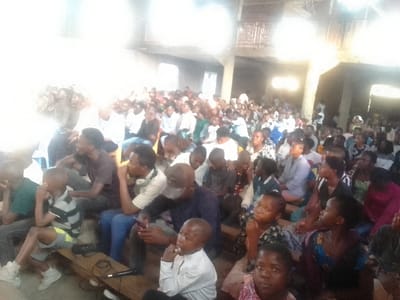 The image depicts an indoor gathering of approximately 100-150 individuals within a large, utilitarian hall in Goma, Democratic Republic of the Congo. The assembly comprises predominantly young adults and adolescents, with children present in the foreground. Individuals are seated on a mix of wooden benches and plastic chairs, arranged in rows, generally facing forward. Observable elements include a young boy in a striped shirt seated on a wooden bench in the lower-left, next to a black microphone with an attached cable. A male subject in the middle-left is holding a mobile phone. The hall features concrete support pillars and an upper-level balcony structure visible in the background. Bright light, originating from the left side of the frame, causes significant overexposure in that area, suggesting an external light source such as a window or doorway.