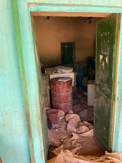 The image is a view from the doorway of a small, cluttered room located in Khartoum, Sudan, at coordinates (latitude: 15.552, longitude: 32.57). The room is sparsely lit, with peeling, ochre-colored walls and a green wooden door on the right. The doorway is framed by a green painted wall.  Inside the room are several large metal drums, a white refrigerator-like box, and a brown plastic basket, all of which are arranged in a haphazard manner. The floor is dusty and littered with crumpled brown paper bags. No people are visible.  A few miscellaneous items, including plastic containers, are scattered on top of the white box.  The room appears to be in disarray and may be used for storage.