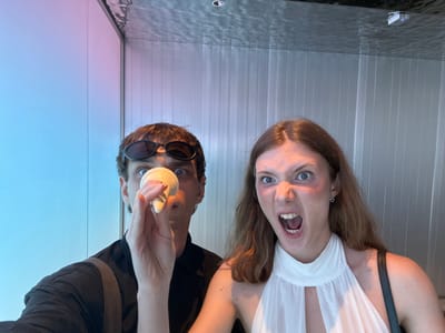 This image captures two individuals, one female and one male, from an upside-down perspective, looking directly down towards the camera.
The female, positioned on the left, has long, light brown hair that appears to float upwards. She wears a white, sleeveless top with a sheer, pleated neckline and a black strap over her shoulder. Her expression is exaggerated, featuring a wide-open mouth revealing teeth and tongue, and wide, startled blue eyes. A small, black heart-shaped earring adorns her left earlobe.
The male, on the right, has short brown hair and is wearing dark sunglasses pushed up on his nose, partially obscuring his eyes. He is dressed in a dark-colored top. His right hand is holding a small, light-colored, possibly cylindrical object or container, which he extends downwards.
The background directly behind their heads features a light gray wall with distinct vertical ridges or panels. Below them, occupying the lower portion of the frame, is a highly reflective surface that shows rippling patterns, suggesting water or a deeply polished, possibly wet, floor. On this reflective surface, a small, silver-grey rectangular device with a blue indicator light and a circular, dark fixture (possibly a drain or floor light) are visible. Further beyond this surface, the wall transitions into a smooth, light-colored gradient, shifting from a subtle pinkish hue to a pale blue, suggesting artificial lighting.
The setting appears to be a modern indoor environment or an art installation, possibly in a contemporary building in Shanghai. The photograph captures a playful and surreal moment, emphasizing the exaggerated expressions and the unusual orientation. No text is visible in the image.