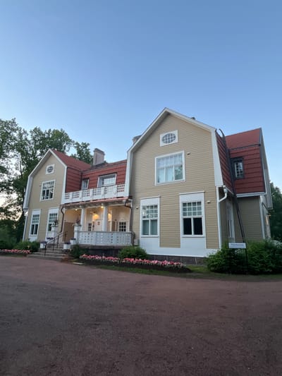 The image shows a large, two-story yellow house with a red tiled roof, located in Vantaa, Finland. The house has multiple windows, some with white frames and others with dark panes. A white porch with decorative railings extends across the front of the house, leading to a set of stairs. Flower beds filled with pink flowers are planted in front of the house. A signpost with Finnish text "Päärakennus" is visible to the right of the house. The house is surrounded by trees and lush green bushes. The sky is clear and blue, suggesting it is either early morning or late evening. The overall scene depicts a well-maintained residential property in a natural setting.