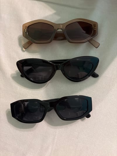 In the city of Voluyak, Bulgaria, three pairs of sunglasses are arranged vertically on a flat, light-colored, subtly textured surface. The topmost pair features translucent brown, geometric-shaped frames with dark lenses and two parallel rectangular metallic accents on each outer temple. The middle pair consists of black cat-eye style frames with dark lenses; partial text "London design" is visible on the right lens. The bottom pair displays thick-rimmed, rectangular black frames with dark lenses, with the word "NEW YORKER" visible on the right lens. All sunglasses are stationary, exhibiting slight reflections on their lenses and casting soft shadows on the surface. No individuals or actions are depicted.
