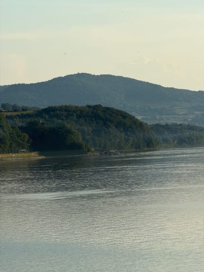 A serene, wide shot captures a tranquil body of water in the foreground, reflecting the soft, pale sky above with subtle ripples on its surface. Beyond the water, a densely forested hill slopes upwards, its trees a deep green against the muted backdrop. In the distance, layers of hazy blue hills rise, suggesting a vast, undulating landscape. The scene is devoid of people or any obvious human activity, emphasizing the natural beauty of the location, possibly a reservoir or lake near Lipie, Poland, known for its picturesque surroundings and natural reserves. The light suggests it is either early morning or late afternoon, casting a gentle glow over the entire scene.