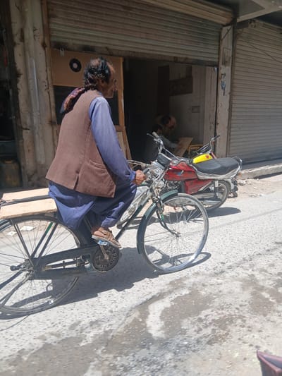 An elderly man with dark hair and a mustache, wearing a blue shirt, a brown vest, and blue shalwar, rides a black bicycle with a wooden plank attached to the rear. He is wearing brown sandals. The man is in motion on a paved street in front of a building with a closed metal shutter. Behind him, a red and yellow motorcycle is parked, and inside the open doorway of the building, two other individuals are visible, one appearing to be working on something. The scene is outdoors, with sunlight casting shadows on the street. The environment appears to be an urban setting, possibly a marketplace or commercial area. This image was captured in Quetta City Tehsil, Pakistan.
