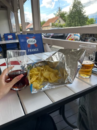 A person's hand with manicured nails holds a glass of dark liquid. Next to the glass is an open bag of potato chips and another glass containing a lighter colored beverage. The setting appears to be an outdoor cafe or restaurant in Velykyi Bychkiv, Ukraine, with tables and chairs visible, and a backdrop of buildings and trees through a window.