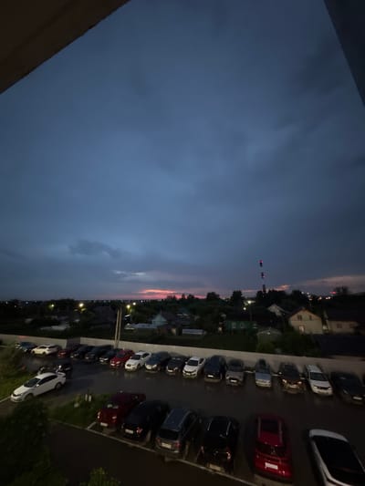 The image shows an aerial view of a parking lot filled with cars, situated in the village of Kondratovo, Russia. The scene is set during twilight, with a dark, cloudy sky. A faint pinkish hue is visible on the horizon, suggesting the sun has recently set. In the background, a cluster of houses with illuminated windows and streetlights can be seen, along with trees and a tall industrial structure with red and white lighting. The parking lot is paved and appears wet, possibly from recent rain. The cars are parked in rows, and include various models and colors, such as white, black, red, and grey. Some of the cars are sedans, while others appear to be SUVs or hatchbacks. The vantage point suggests the photo was taken from a higher elevation, possibly a building or balcony overlooking the parking lot and surrounding area.
