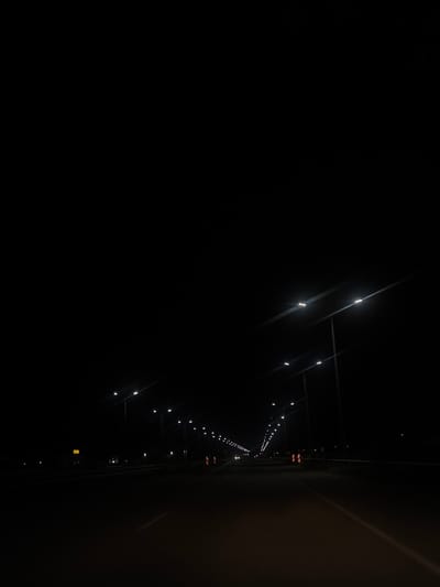 The image depicts a street lit by streetlights at night, with a dark sky. The perspective suggests a view from a moving vehicle on a road, as indicated by the lines on the road surface and the angled streetlights receding into the distance. The streetlights are modern, with multiple LED bulbs. Orange and white striped markers are visible on the side of the road, suggesting a highway or major road. In the distance, other vehicles can be seen. The overall scene is one of a dimly lit road at night, likely in an urban or suburban area. The location is specified as Asenovgrad, Bulgaria.