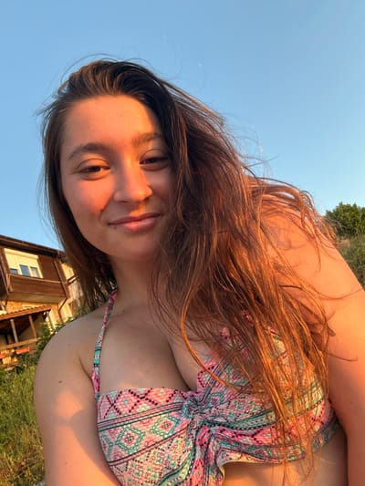 A young woman with long, brown hair is taking a selfie outdoors. She is wearing a colorful bikini top with a geometric pattern. The background shows a wooden building, green grass, and a clear blue sky, suggesting a coastal location. The lighting indicates it is either sunrise or sunset, casting a warm glow on her face and hair. The image appears to have been taken in Sozopol, Bulgaria, a city known for its beaches and historic architecture.