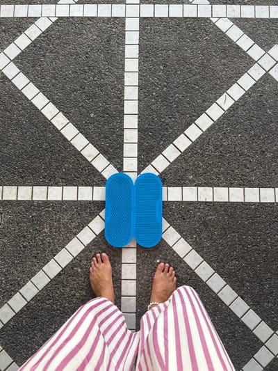 A top-down view captures a person's feet and lower legs standing on a patterned asphalt surface. The ground is a mosaic of dark grey asphalt interspersed with a geometric pattern of white square tiles, creating intersecting diagonal and horizontal lines that converge in the center. Placed precisely in the center of this pattern are two bright blue, oval-shaped objects. These objects appear to be yoga or acupuressure mats, with a textured, dimpled surface and a visible imprint of text on the top one that reads "SADHU BOARD". The person is wearing loose-fitting, wide-legged pants with bold horizontal stripes in white and light purple. One foot is positioned to the left of the blue mats, and the other is to the right, close to the edge of the frame and the striped pants. The lighting suggests it is daytime, likely outdoors, given the texture of the asphalt. The setting appears to be an outdoor public space, possibly a walkway or plaza, in Ratsada, Thailand. The overall impression is a snapshot of everyday life, perhaps a moment of pause or preparation for an activity.