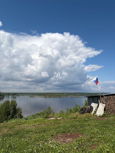 This landscape image, taken in Olikhovka, Russia, presents a serene outdoor scene under a bright sky. The upper left portion of the frame is dominated by a clear blue sky, partially filled with immense, fluffy white cumulus clouds. These clouds extend across the middle of the sky, eventually giving way to thinner, less distinct cloud formations towards the right.
A wide, placid body of water, likely a river or lake, stretches across the mid-ground, reflecting the light from the sky. Its distant bank is lined with a dense strip of green trees and vegetation, with faint hints of human structures nestled within the foliage.
In the foreground on the right, a vibrant green grassy area unfolds, dotted with numerous white dandelion seed heads and a few yellow dandelion blooms. A dirt path or bare ground is visible traversing this area. Further right and slightly elevated, there's a rustic structure with a dark, corrugated wall, accompanied by a large stack of cut firewood. Several old tires are placed near this structure, along with some white rectangular objects. A white flagpole extends upwards from this area, bearing a Russian flag, distinguished by its horizontal white, blue, and red stripes, gently fluttering in the breeze.
The scene depicts a peaceful daytime, likely late morning or early afternoon, with abundant sunlight and fair weather, characterized by the bright clouds and clear blue sky. No specific activities or individuals are visible, and there is no discernible text in the image.