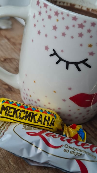The image shows a white mug with pink stars and a face design, placed on a wooden surface. In front of the mug are two candy bars. One is yellow with the word "МЕКСИКАНА" in black lettering, and the other is white with red lettering that appears to read "Rafaello" followed by Russian text. The scene appears to be indoors, possibly in a home or cafe setting, given the presence of a mug and candy. The lighting suggests a natural light source. There are no people visible in the image. The overall impression is a still life composition of a beverage and treats. It is impossible to determine the specific location in Забалоцкі сельскі Савет, Belarus from the provided image.