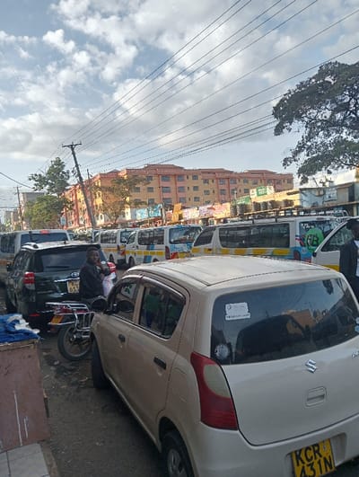 An urban scene depicts a street filled with various vehicles. A beige Suzuki car is parked prominently in the foreground with the number plate KCR 431N. Behind it, a motorcycle and a dark SUV are visible. A man stands near the SUV. In the background, a row of minibuses and buses line the street, against a backdrop of a large building and a sky dotted with clouds and power lines. The scene seems to capture a typical busy day in an urban environment.