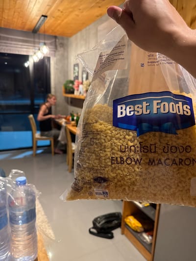 In Rawai, Thailand, a person's hand holds a clear plastic bag filled with elbow macaroni pasta. The bag displays the "Best Foods" logo and text in English and Thai, indicating "ELBOW MACARON." The background is blurred but suggests an indoor setting with wooden ceiling beams and lighting fixtures. Further in the background, a woman is seated at a table, appearing to be eating, with chairs and other objects visible. Bottles of water are present in the foreground.