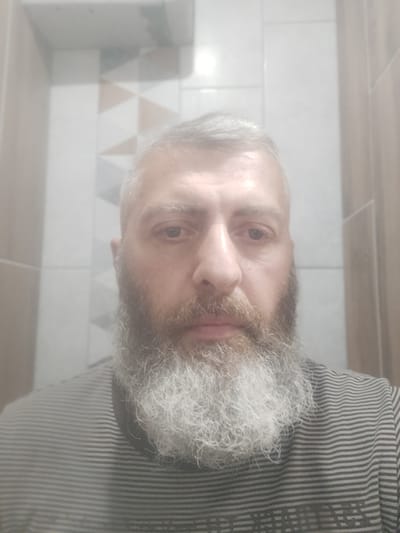 A medium shot, frontal, waist-up photograph shows a man with grey hair and a full grey beard. He is wearing a grey and black striped shirt. The background appears to be a bathroom with tiled walls. The man's expression is neutral. The image is slightly blurry and taken in dim lighting, likely indoors, possibly in Perepilchyntsi, Ukraine.