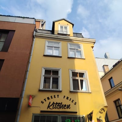 The image shows a low-angle shot of the exterior of a yellow building in Riga, Latvia, under a bright, partly cloudy sky. The building features multiple windows with white frames. A sign above a doorway reads "STREET FRIES Kitchen" in black lettering. To the left of the yellow building is a darker brown building, and to the right, a lighter peach-colored building is partially visible. There are no people visible in the image.