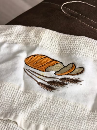 The image shows an embroidered image of a loaf of bread with slices and wheat stalks on a white towel. The towel appears to be on a brown surface, possibly a piece of furniture. There are no people or actions depicted in the image. The image does not contain any information that would allow for its location in Sterlitamak, Russia to be determined.