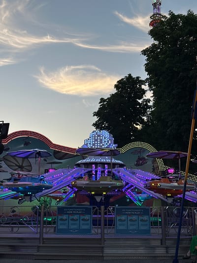 The image shows a close-up of a carnival ride at dusk, likely in Vienna, Austria. The ride is illuminated with many colorful lights, primarily purple and white, with some red accents. The ride appears to be a spinning amusement ride with multiple cars or gondolas.
In the background, tall trees with dark green foliage are visible, suggesting an outdoor setting, possibly a park or fairground. The sky is a pale blue with wispy, elongated clouds that are illuminated by the setting sun, creating a soft, warm glow.
One of the ride's components features a sign with German and English text. The visible text on the sign reads "SO FUNKTIONIERT'S - HOW TO PLAY -" followed by three illustrated steps showing how to operate the ride. The illustrations depict a person in a car-like vehicle on the ride.
The overall impression is one of a vibrant and exciting evening at a fair, with the lights of the ride contrasting with the darkening sky and lush greenery. The image is taken at an angle, giving a dynamic perspective of the amusement park attraction.