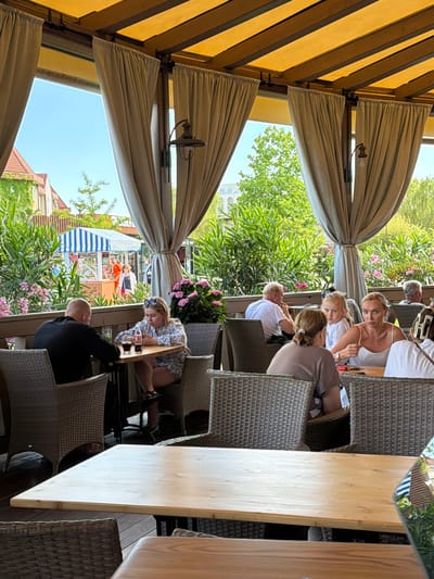 In the city of Sirius, Russia, a group of people are seated at tables on an outdoor patio of a restaurant. The patio is covered by a yellow awning with wooden beams, and draped with light-colored curtains that partially obscure the view of the outside. In the foreground, two people, a man in a black shirt and a woman in a patterned top, are seated at a table, with drinks and cell phones on the table. In the middle ground, a woman with blonde hair and a white top is talking to a young girl, while another woman with brown hair in a grey t-shirt sits with her back to the camera. Further back, several other individuals are visible at tables. The background shows greenery, buildings, and a striped canopy of a stall. The atmosphere appears relaxed and social, with people engaged in conversation and enjoying their time outdoors.