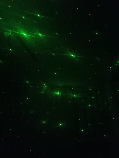 The image shows a dark room illuminated by a green laser light projector, creating a starry night effect on the ceiling and walls. The green lights are scattered across the surface, some appearing as small dots while others have a more diffused glow with visible light rays. There is no visible evidence of people, furniture, or any specific location markers that would indicate it is in the city of Bălți, Moldova. The focus is solely on the pattern of the projected green lights against a dark background.