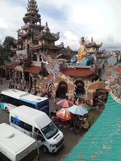 The image shows a large, ornately decorated temple complex in Xuân Thành, Vietnam. The architecture is characterized by intricate dragon motifs, numerous tiered roofs, and elaborate ceramic decorations. In the foreground, two buses and a white minivan are parked on a paved area. People are seen walking around the temple grounds and near the vehicles, some of whom appear to be tourists. Several stalls with colorful umbrellas are set up, suggesting a market or vendor area. The sky is partly cloudy, and the overall impression is one of a vibrant cultural and tourist destination.