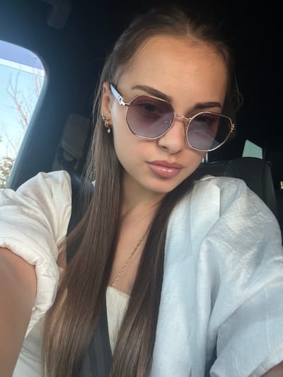 A young woman with long, brown hair, wearing sunglasses, a white top, and a white jacket, is sitting in the driver's seat of a car. She is taking a selfie, with her left arm visible in the foreground. The background shows the interior of the car, including a black seat and a window with a glimpse of trees and sky. The image is taken outdoors, suggesting a sunny day. The location is specified as Lozen, Bulgaria.