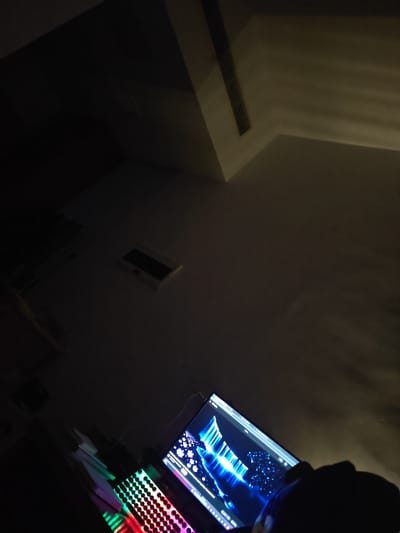 A dimly lit room in Jiangning District, China, features a computer setup with a monitor displaying a vibrant blue and white abstract image. In the foreground, a mechanical keyboard with colorful RGB backlighting is visible, suggesting a gaming or professional setup. The overall ambiance is dark, with the computer's illumination providing the primary light source. The ceiling shows a ventilation grate, and the walls are bare.