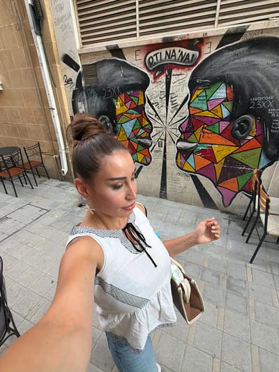 A woman is captured in a selfie, standing in an outdoor urban setting in Nicosia, Cyprus. She has her hair pulled back in a bun and is wearing a white top with black trim and blue jeans. Her right arm is raised, and she is looking slightly off-camera with a pleasant expression. Behind her, a vibrant mural adorns a wall, featuring two stylized faces filled with colorful geometric shapes. The ground is paved with gray flagstones. The scene appears to be a public space, possibly a street or courtyard, with some tables and chairs visible in the background, suggesting a cafe or restaurant area. The lighting and shadow play indicate it is daytime. The mural itself is a prominent feature, with visible text in Greek and possibly other languages. Some of the legible text on the mural includes "OTI NA 'NAL," "23XX," "ΑΝΘΟΥΠΟΛΗ," and what appear to be signatures or tags. The overall mood is casual and relaxed, with the woman seemingly enjoying her surroundings.