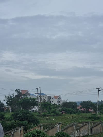 The image depicts a hazy, overcast sky over a semi-urban landscape, possibly in Rafin Par, Nigeria. In the middle ground, a cluster of multi-story buildings with white facades and various roof colors, including orange and blue, are visible amongst lush green trees. Some of these buildings appear to be under construction or recently built, with visible windows and architectural features. Electrical poles with wires crisscross the scene, indicating utility infrastructure. To the left, a small, red and yellow auto-rickshaw is parked in a grassy area. The foreground is dominated by a weathered, low-lying concrete structure with a stepped top, overgrown with green vegetation. The overall impression is of a developing area with a mix of residential buildings and natural greenery under a diffused light.