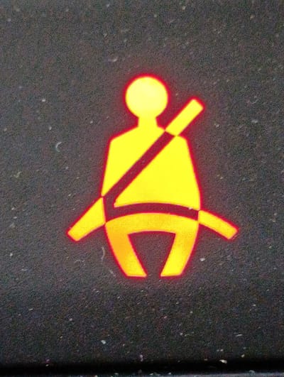The image shows a close-up of a yellow seatbelt reminder icon illuminated against a dark, textured surface, likely a dashboard. The icon depicts a stylized human figure wearing a seatbelt. There are no people or specific city locations visible in the image. The surface appears to be a car's interior.
