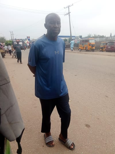 A medium-shot, eye-level outdoor photograph captures a Black man in a blue t-shirt and black pants standing on a dirt road in Kubwa, Nigeria. He faces forward with his arms behind his back and his head slightly turned to the right. The man has a dark complexion and a shaved head. He is wearing gray and white striped sandals.
The background is a busy street scene. To the left, a market is in progress with people carrying produce and other goods. A cart loaded with green leafy vegetables is visible. In the mid-ground, several yellow auto-rickshaws, commonly known as "Keke Napep," are parked on the side of the road. Further back, a red car is also visible. A tall utility pole with wires stretches across the top of the frame. The sky is overcast, suggesting a cloudy day. The ground is a mixture of dirt and gravel with scattered debris. A portion of a light-colored object, possibly a vehicle or a tent, is in the foreground on the left, obscuring part of the scene. The overall impression is a typical day in a Nigerian urban market environment.