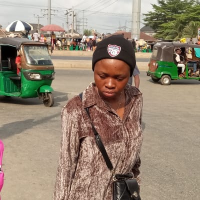 A person wearing a black beanie and brown patterned shirt stands on a street. Several green and black motorized tricycles, commonly known as "keke," are visible around her. A black handbag with a strap hangs on the person's shoulder. The background is a bustling street scene with people, power lines, and various buildings. The lighting indicates a sunny day.