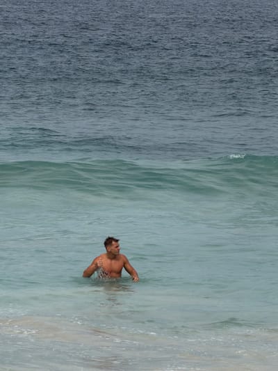 A shirtless man is waist-deep in the ocean water, facing right, with a thumb raised in a gesture of approval. He has short, dark hair and is tanned. The water is turquoise and appears to be shallow near the shore where there are white-capped waves breaking. The ocean extends to the horizon, with darker, choppier water visible in the distance. The scene is set on a beach in Manggis, Indonesia.