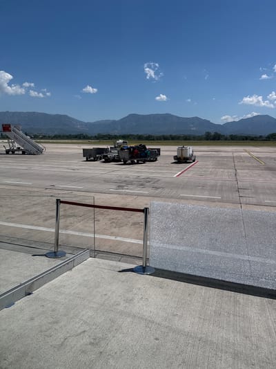 The image depicts an outdoor scene at an airport, likely at Tirana International Airport Nënë Tereza (TIA) in Kruja, Albania, given the "TIA" visible on the airstair. The foreground shows a concrete surface with a metal stanchion and red velvet rope barrier, suggesting a waiting or queuing area. A textured, possibly reflective, surface is visible to the right of the barrier. The mid-ground of the image features the airport tarmac, with various pieces of ground support equipment.
Prominently displayed on the left is a white airstair, labeled "TIA" and "ALBSIG." To the right of the airstair are several baggage carts loaded with luggage. Further to the right, a small white service vehicle is parked on the tarmac. The tarmac is marked with white and yellow lines, indicating taxiways or runways.
In the background, a grassy area leads to a line of trees, beyond which rise a range of mountainous terrain. The sky is a clear, bright blue with scattered white cumulus clouds. The overall impression is one of a functional airport environment under a sunny, clear sky, with the mountains of Albania providing a scenic backdrop. There are no people visible in the image.