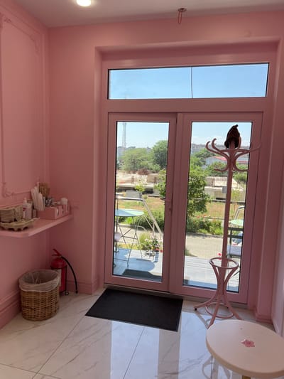 This image shows an interior scene, likely a cafe or shop, with pink walls and white marble-tiled flooring. A large glass door with two panels looks out onto an outdoor patio area. To the left of the door, a pink shelf holds various items, including what appear to be disposable cups, napkins, and utensils in pink organizers, as well as a fire extinguisher in a stand. A woven basket trash can is positioned on the floor next to the shelf. In front of the door, a dark mat is laid on the floor. A pink coat rack stands to the right of the door, with a dark hat placed on top. In the foreground, a small, round, pink table has a pink spill on its surface. The outdoor view through the glass door reveals a patio with green chairs and tables, some potted plants, and a partially constructed building or scaffolding in the background. The sky is clear and blue. The scene is located in the city of Odesa, Ukraine.