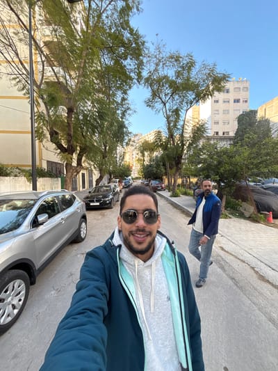 A selfie-style photograph captures two adult males on a paved city street in Tangier, Morocco, under a clear blue sky.
In the immediate foreground, facing the camera and positioned slightly left of center, is an adult male with dark hair, a beard, and dark sunglasses. He is smiling and wears a dark teal puffer jacket over a light grey hooded sweatshirt with light blue trim. His upper torso and head are visible.
Behind and to the right of the foreground subject, on the sidewalk, another adult male is walking towards the camera. He has dark hair and a beard, and is wearing a blue and black puffer jacket over a white sweater and blue jeans.
The street, which appears to be residential or mixed-use, runs into the background, lined with various parked vehicles. A silver SUV is partially visible on the left, next to the foreground subject. Further down the street, a black BMW sedan and other cars are parked on the right side.
Buildings of varying heights and architectural styles line both sides of the street. On the left, a multi-story building with cream-colored walls and yellow horizontal trim is visible. On the right, a taller, plainer white building with numerous windows and satellite dishes on its rooftop is visible in the distance. The street is also lined with numerous mature trees featuring green foliage. On the right sidewalk, near the walking individual, there is a black tarp covering an object, with a small orange cone nearby. A dark street lamp post is visible on the far left. The lighting suggests daytime conditions with a clear sky.