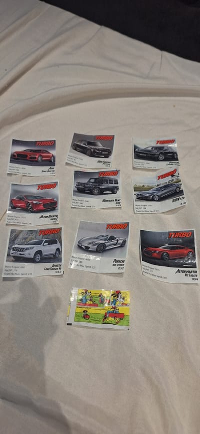 The image shows a collection of "Turbo" brand chewing gum wrappers scattered on a light-colored surface, likely a bed sheet. The wrappers feature different car models and their specifications, including engine size, horsepower, and maximum speed. Some of the car models include Aston Martin V12 Zagato (094), Porsche 918 Spyder (052), Toyota Land Cruiser V8 (032), Mercedes Benz G500 (019), BMW 520 (012), Audi Sport Quattro (125) and others. One wrapper features cartoon characters. The wrappers are slightly wrinkled and out of alignment. The background is a simple, slightly wrinkled bed sheet with the edge of something dark on the left, out of focus. There is no information in the image regarding the location as Pernik, Bulgaria.
