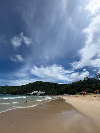 The image depicts a wide, sandy beach in Rawai, Thailand, under a partly cloudy sky with dramatic cloud formations. Gentle waves lap the shore, creating a foamy edge on the wet sand. In the shallow water, a single person is partially submerged, appearing to relax or swim. The beach is lined with lounge chairs and umbrellas, suggesting a resort area. Red and yellow flags, possibly indicating a designated swimming area or surf rescue station, are visible on the sand. Behind the beach, a lush, green, tree-covered hill rises, with a cluster of white buildings visible partway up its slope. The overall scene conveys a peaceful and scenic beach environment on a bright day.