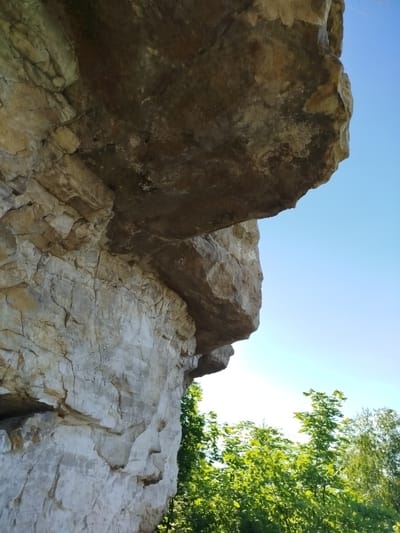 An overhanging rock formation is captured in this low-angle outdoor shot. The rock is light-colored and appears to be made of layered stone, with visible cracks and textures. Above the rock, a clear, bright blue sky fills the upper portion of the frame. Below and to the right of the rock, a dense canopy of vibrant green trees with lush foliage is visible. The image does not depict any people, actions, or interactions. The location is not specified beyond general environmental features.