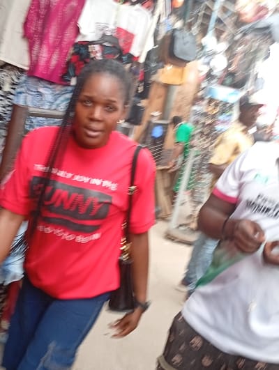 A woman in a red t-shirt and blue jeans stands in a crowded marketplace. The shirt has the word "FUNNY" printed on it. She has dark hair and a shoulder bag. Behind her, clothing racks and other merchandise are visible. Other people are milling about in the background. The image is somewhat blurry.