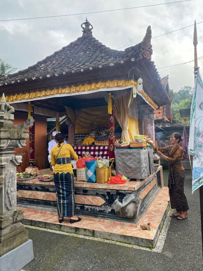 The image depicts a religious ceremony at a temple, likely in Gianyar, Indonesia. A woman in a brown traditional outfit is lighting incense sticks, with smoke rising. Another woman in a vibrant yellow and patterned sarong stands facing a shrine, arranging offerings. The shrine features a tiered platform decorated with tiles, holding various items including woven baskets, food items, and offerings. The temple structure has a tiled roof adorned with decorative elements and yellow fabric draped from the eaves. In the background, there are trees and other structures, suggesting a rural or semi-urban setting. A flag with intricate designs is visible on the right side of the frame. The overall scene conveys a sense of cultural and spiritual observance.