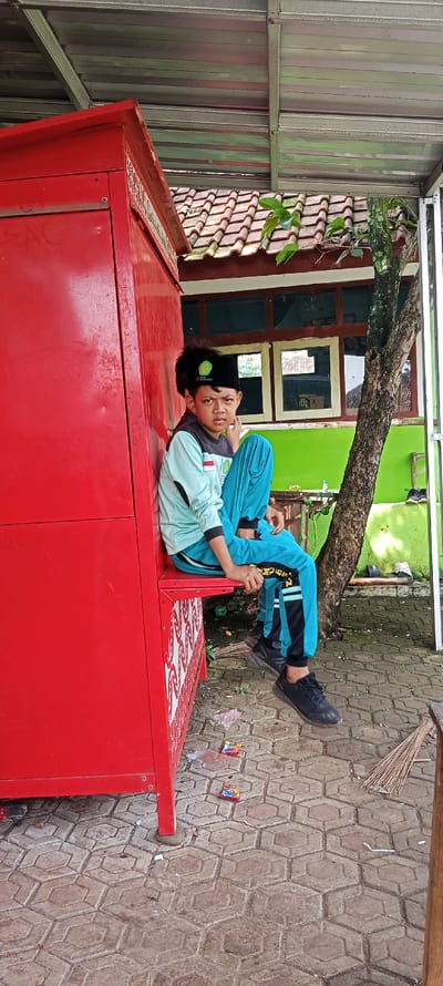 The scene takes place in Cangkuang, Indonesia, at the coordinates -7.065, 107.551. A young boy, wearing a turquoise shirt, blue and black striped pants, and a black cap with a green symbol, sits on a red wooden structure with decorative carvings. He is leaning to the right, looking to the left, and resting his arm on the structure. His legs are extended forward, and his right foot is resting on the ground. The scene is in front of a red-bricked building with a green wall, and a large tree behind it. The ground is made of hexagonal-shaped tiles. There are a few items of trash on the ground, including a piece of plastic, a few cigarette packets, and a small, thin broom. The scene is well-lit and captures a moment of quiet reflection.