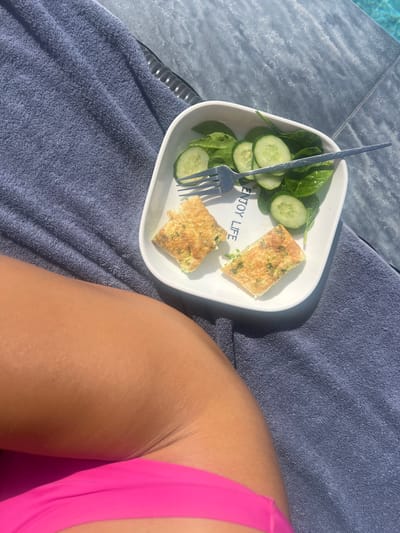 A person's tanned leg and hip, clad in bright pink swimwear, rests on a textured blue towel. Beside the leg, a white rectangular plate holds a light meal consisting of two folded omelet pieces with flecks of green herbs, a side salad of spinach leaves, and sliced cucumbers. A silver fork is placed on the plate, with its tines resting amongst the salad. The setting appears to be outdoors, with a tiled surface suggesting a poolside or patio area, and a hint of blue water in the upper right corner, indicative of a swimming pool. The overall scene suggests a relaxed moment, possibly a healthy breakfast or snack by the water. The location is identified as Baan Bang Rak, Thailand.